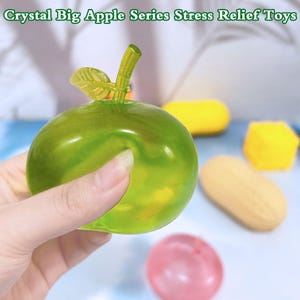 Puede incluir: Un juguete antiestrés translúcido en forma de manzana verde con tallo y hoja. El texto "Crystal Big Apple Series Stress Relief Toys" está en la parte superior. Otros juguetes antiestrés coloridos están en el fondo, incluyendo un cubo amarillo y una bola rosa.