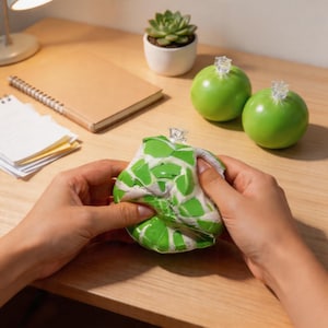 Può includere: Una persona sta stringendo una palla antistress con un motivo a mosaico verde e bianco. Due sfere decorative verdi con cime trasparenti sono vicine. Una piccola succulenta in un vaso bianco e un taccuino sono anche sulla superficie di legno.