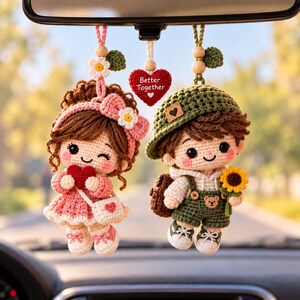 May include: Two crocheted dolls hanging from a rearview mirror. The left doll wears a pink dress, a matching headband with a white flower, and carries a red heart. The right doll wears a green hat, green overalls with a bear button, and holds a sunflower. A red heart charm with the text Better Together hangs between them.