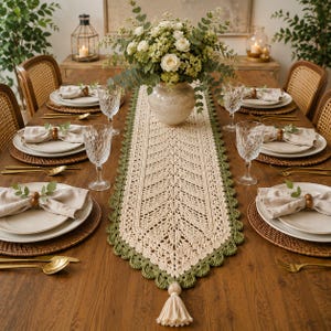 May include: A long, rectangular crochet table runner in cream with a scalloped sage green border and a cream tassel at the end. The runner features an intricate leaf-like pattern and is centered on a wooden table with place settings.