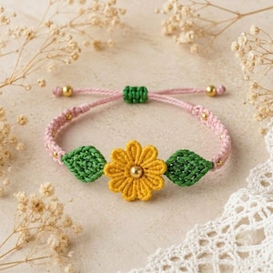 May include: A macrame bracelet featuring a central yellow flower with a gold bead centre, flanked by two green leaves. The band is made of light pink cord with small gold beads integrated into the weave and a green adjustable sliding knot closure.