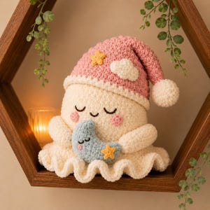 Op de afbeelding: Een crèmekleurige gehaakte knuffel met een slaperige uitdrukking en roze wangen, met een roze slaapmuts versierd met een gele ster en een witte wolk. De knuffel houdt een kleine blauwe halve maan vast met een gele ster eraan bevestigd.