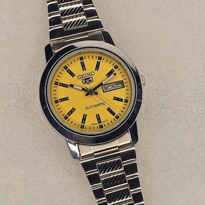 May include: A silver Seiko automatic wristwatch with a vibrant yellow face. The watch has a day and date display and a silver link strap. The watch face has black hour markers and hands. The watch is marked "SEIKO" and "AUTOMATIC".