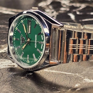 May include: A silver-toned wristwatch with a green face. The watch has a stainless steel band and a date display. The watch face features the brand name "Seiko" and the word "Automatic". The watch is displayed on a textured surface.