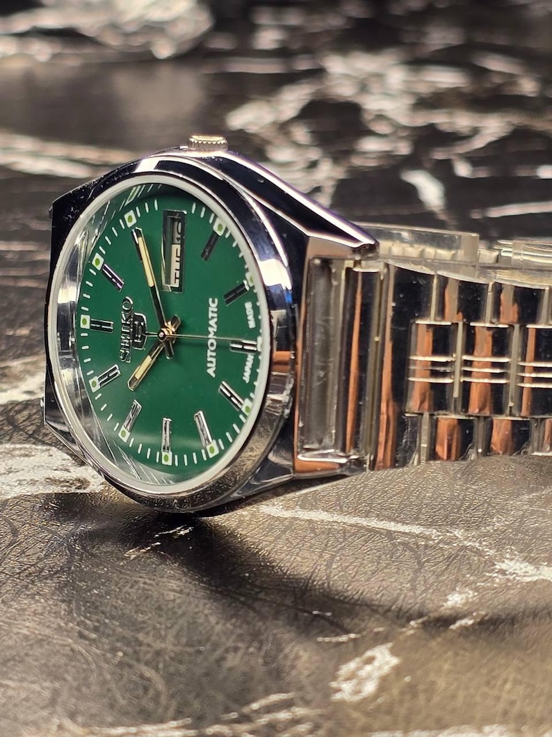 May include: A silver-toned wristwatch with a green face. The watch has a stainless steel band and a date display. The watch face features the brand name "Seiko" and the word "Automatic". The watch is displayed on a textured surface.