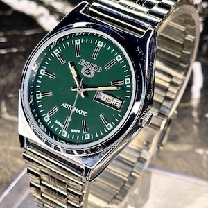 Peut inclure: Une montre-bracelet automatique Seiko 5 en acier inoxydable argenté avec un cadran rond vert, des index argentés, des aiguilles luminescentes et un guichet jour-date à trois heures. La montre possède un bracelet à maillons en métal.