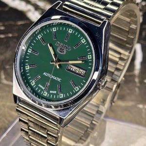 Op de afbeelding: Een roestvrijstalen Seiko 5 automatisch horloge met een groene wijzerplaat. Het horloge heeft een dag- en datumweergave en een zilverkleurige metalen band. De wijzerplaat heeft zilveren uurmarkeringen en wijzers.