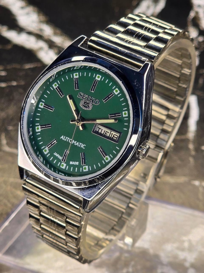 May include: A stainless steel Seiko 5 automatic watch with a green dial. The watch features a day and date display, and a silver-tone metal band. The watch face has silver hour markers and hands.
