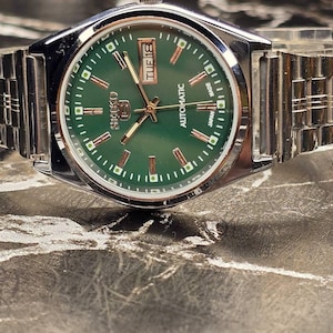 May include: A silver-toned Seiko automatic wristwatch with a green dial. The watch face displays the day and date, with gold-colored hands and markers. The watch has a stainless steel band.