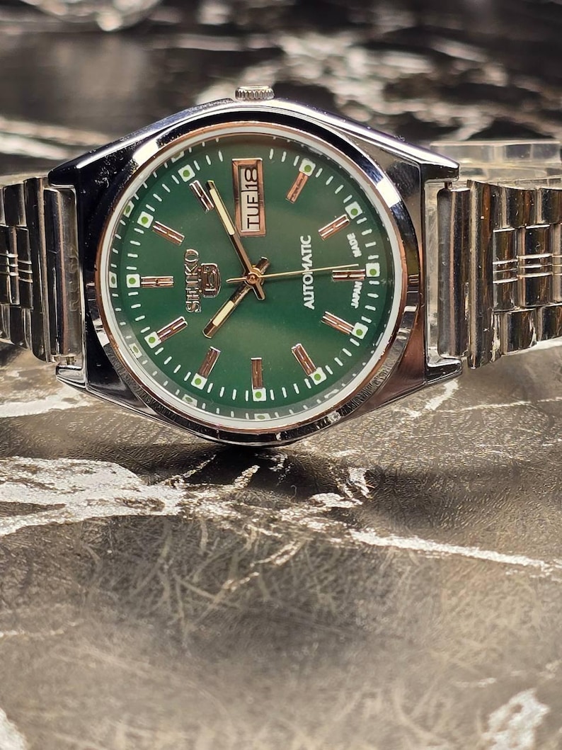 May include: A silver-toned Seiko automatic wristwatch with a green dial. The watch face displays the day and date, with gold-colored hands and markers. The watch has a stainless steel band.