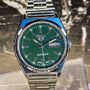 May include: A silver-toned Seiko 5 automatic wristwatch with a green dial. The watch features a day and date display, luminous hands and markers, and a stainless steel band. The watch face displays the text "SEIKO 5 AUTOMATIC JAPAN MADE".