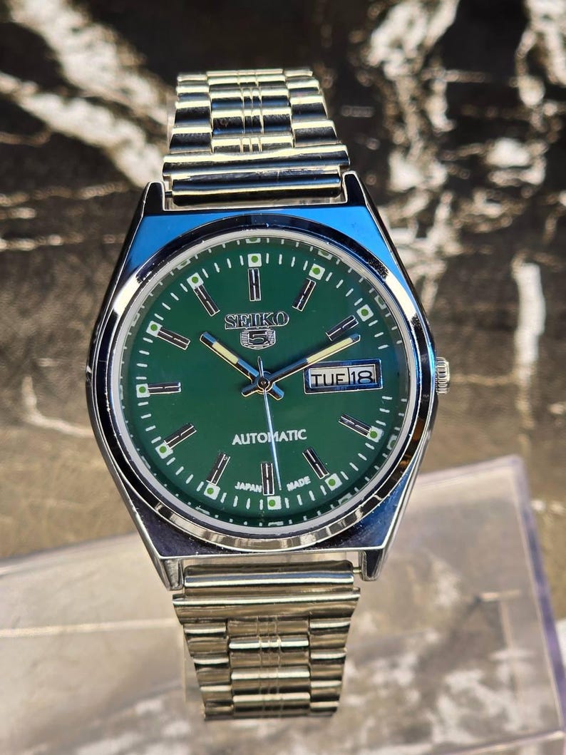 May include: A silver-toned Seiko 5 automatic wristwatch with a green dial. The watch features a day and date display, luminous hands and markers, and a stainless steel band. The watch face displays the text "SEIKO 5 AUTOMATIC JAPAN MADE".