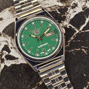 May include: A silver-toned Seiko 5 automatic watch with a green dial. The watch face displays the day and date, with a silver band. The watch is marked "Japan Made".