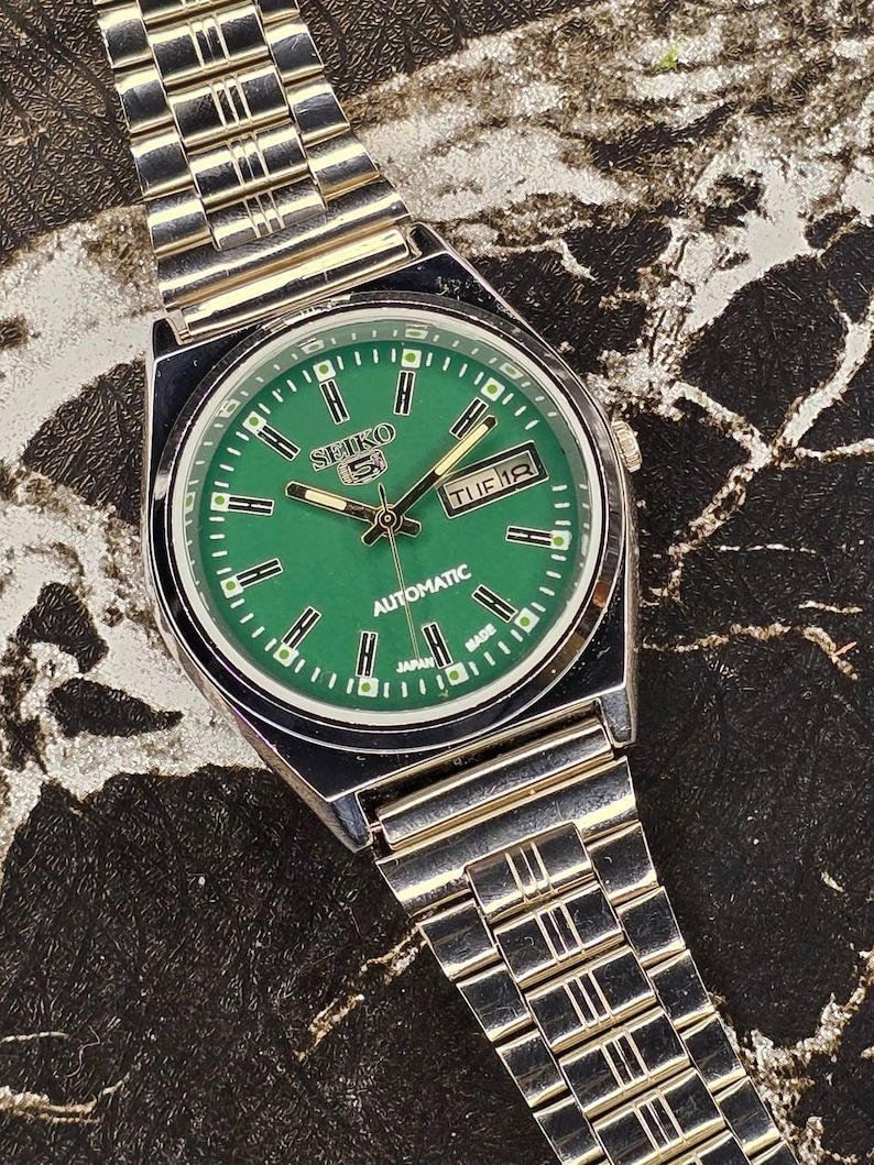May include: A silver-toned Seiko 5 automatic watch with a green dial. The watch face displays the day and date, with a silver band. The watch is marked "Japan Made".