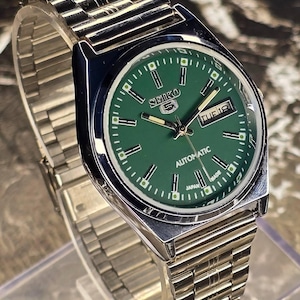 May include: A silver-toned Seiko 5 automatic wristwatch with a green dial. The watch features a day and date display, and a silver-toned metal band. The watch face has white hour markers and hands.