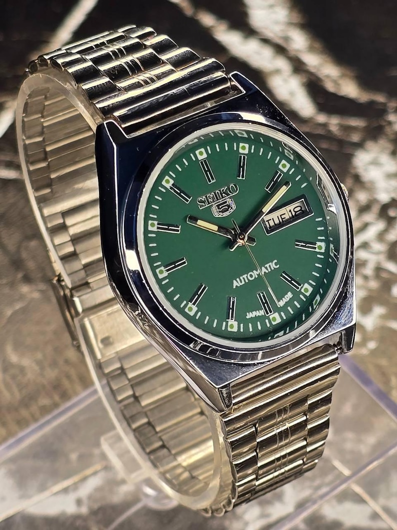 May include: A silver-toned Seiko 5 automatic wristwatch with a green dial. The watch features a day and date display, and a silver-toned metal band. The watch face has white hour markers and hands.