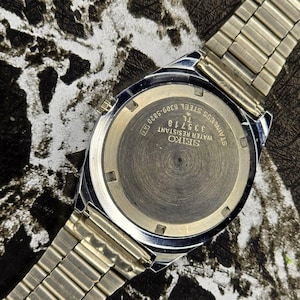 May include: A close-up of a silver-toned Seiko watch, showcasing the watch back and a portion of the metal band. The watch face displays the text "SEIKO WATER RESISTANT STAINLESS STEEL 335718 TL 6309-5820 A[T]".