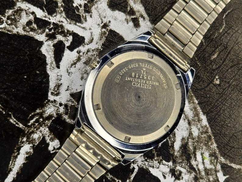 May include: A close-up of a silver-toned Seiko watch, showcasing the watch back and a portion of the metal band. The watch face displays the text "SEIKO WATER RESISTANT STAINLESS STEEL 335718 TL 6309-5820 A[T]".