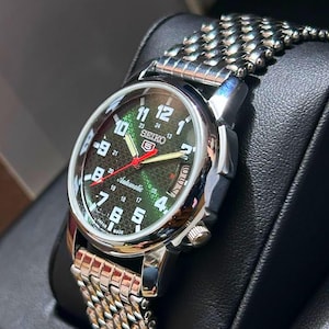 May include: A Seiko 5 automatic wristwatch with a silver case and a textured green dial. The watch has black numerals, a red second hand, and a silver and gold metal band. The watch is displayed on a black surface.