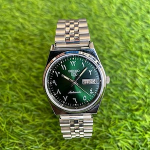 May include: A silver-tone stainless steel Seiko 5 automatic wristwatch featuring a sunburst emerald green dial with Eastern Arabic numerals, silver hands, and a day-date window at the three o'clock position, finished with a metal link bracelet.