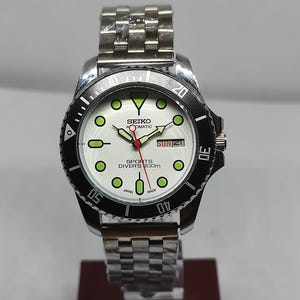 Pode incluir: Relógio de mergulho automático Seiko prateado com moldura giratória preta. O mostrador branco apresenta marcadores circulares verdes, exibição de dia e data e ponteiro de segundos vermelho. Pulseira de aço inoxidável.