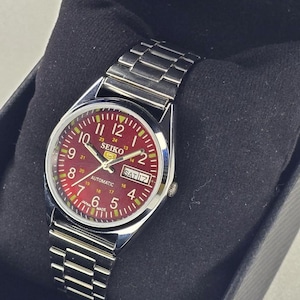 May include: A stainless steel Seiko automatic wristwatch with a red dial. The watch features a day and date display, luminous hands, and a stainless steel band. The watch is presented in a black box.