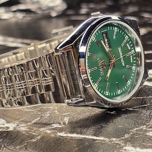 May include: A stainless steel wristwatch with a green dial. The watch has a silver band and a date display. The watch face has gold-colored hands and markers. The watch is labeled "Seiko Automatic Japan".