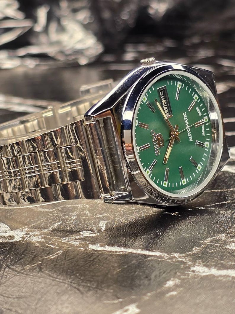 May include: A stainless steel wristwatch with a green dial. The watch has a silver band and a date display. The watch face has gold-colored hands and markers. The watch is labeled "Seiko Automatic Japan".