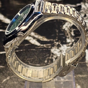 May include: A silver-toned wristwatch with a green face and a stainless steel band. The watch has a rectangular case with a dark blue bezel. The band is composed of small, connected metal links. The watch is displayed on a clear plastic stand.
