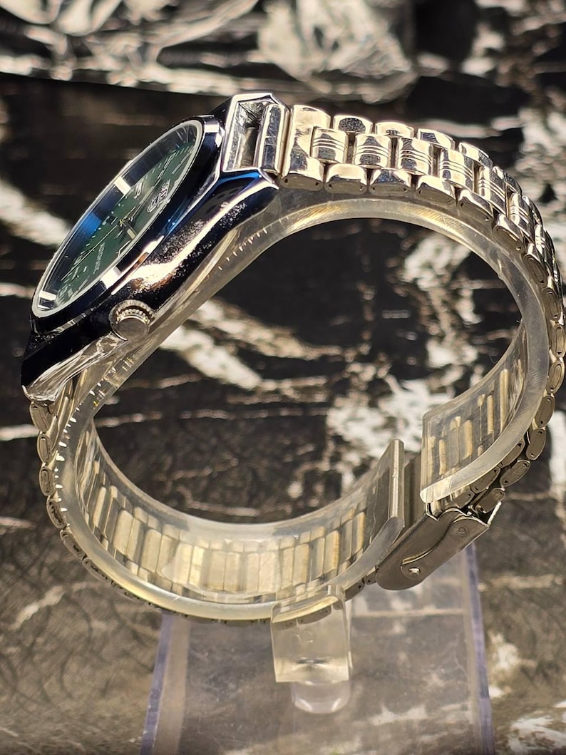May include: A silver-toned wristwatch with a green face and a stainless steel band. The watch has a rectangular case with a dark blue bezel. The band is composed of small, connected metal links. The watch is displayed on a clear plastic stand.