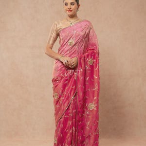 Puede incluir: Un sari de color rosa vibrante con intrincados bordados florales dorados y lentejuelas en toda la tela. La prenda se combina con una blusa de color dorado metálico de manga corta. La palabra yuvti está impresa en blanco sobre el sari.
