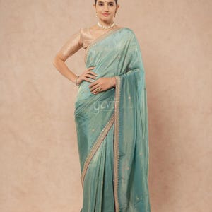 Puede incluir: Un sari de color verde azulado con un brillo sutil y un delicado bordado a lo largo del borde. La prenda se combina con una blusa de color beige claro de manga corta. La tela muestra pequeños motivos florales dispersos por el cuerpo.
