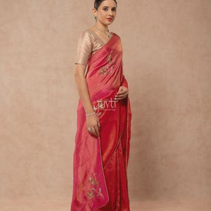 Puede incluir: Un sari de color rojo y rosa vibrante con un brillo dorado metálico, combinado con una blusa beige de manga corta. El sari está adornado con delicados bordados florales en hilo verde y dorado, y el texto "ravti" es visible en la tela.