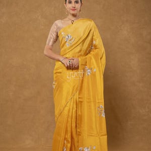 Puede incluir: Un sari de seda amarillo mostaza vibrante con delicados bordados florales plateados dispersos por la tela. La prenda incluye una blusa a juego de manga corta en un tono beige más claro, con la palabra yuvti impresa en blanco sobre la tela.