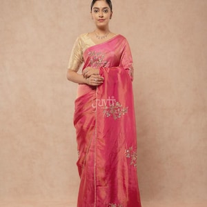 Puede incluir: Un sari de color rosa vibrante con un brillo sutil y delicados bordados florales en hilo dorado y plateado. La prenda se combina con una blusa dorada texturizada de manga corta. La tela cae con elegancia, mostrando intrincados motivos florales.