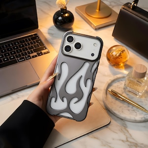 May include: A gray and white flame design phone case. The case is held in a person's hand. The phone case has a black border and a gray background with white flame accents. The phone case is on a white surface.