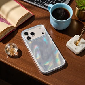 Puede incluir: Una funda transparente e iridiscente para teléfono con un borde azul claro descansa sobre una superficie de madera. La funda tiene un efecto holográfico. Una taza azul claro con café, un bolígrafo y una esfera decorativa de vidrio con copos de oro también están sobre la mesa.