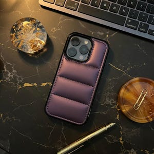 May include: A plum-colored, quilted phone case with a black border rests on a dark, marbled surface. The case has a stitched, padded design. A gold pen, a clear resin coaster with gold flakes, and a small, round, amber-colored dish with a paper clip are also visible.