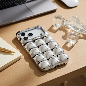 Op de afbeelding: Een glanzende, zilveren telefoonhoes met een uniek, gestructureerd ontwerp. De hoes heeft een reeks afgeronde, belachtige vormen over het oppervlak. De telefoonhoes ligt op een lichtgekleurd houten oppervlak, met een laptop en andere objecten op de achtergrond.