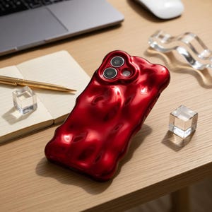 May include: A glossy, red phone case with a unique, textured design. The case features three camera cutouts and a flowing, organic shape. The case is on a wooden surface, with a notebook, pen, and other decorative items in the background.