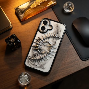 May include: A smartphone case with a white and black marbled design and a silver, stylized, spiky design. The case has a black border and is lying on a wooden surface, with other decorative objects.