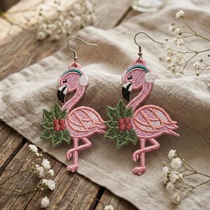 Puede incluir: Un par de pendientes de flamenco rosa con picos negros, hojas verdes y bayas rojas. Los flamencos tienen orejeras y están bordados con un patrón de puntada detallado. Los pendientes se exhiben sobre una tela de color neutro.