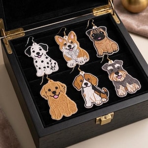 Könnte beinhalten: Sechs bestickte Ohrringe in Hundeform, die einen Dalmatiner, einen Corgi, einen Mops, einen Golden Retriever, einen Beagle und einen Schnauzer zeigen. Jeder Ohrring hat einen goldfarbenen Metallhaken und liegt in einer schwarzen Schmuckschatulle.