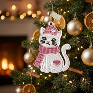 Op de afbeelding: Geborduurde witte kattenkerstversiering, met een roze hoed en sjaal, met een hart op de borst. De versiering hangt aan een kerstboom met gouden ballen en gedroogde sinaasappelschijfjes.