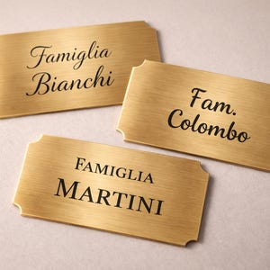 May include: Three rectangular brass nameplates with rounded corners are displayed. Each plate has black text: "Famiglia Bianchi", "Fam. Colombo", and "FAMIGLIA MARTINI". The plates are on a light-colored surface.