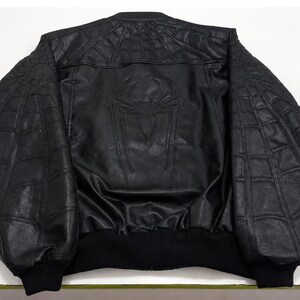 K&ouml;nnte beinhalten: Eine schwarze Bomberjacke mit einem strukturierten Spinnennetz-Muster, das auf die Schultern und &Auml;rmel gepr&auml;gt ist. Auf dem R&uuml;ckenteil befindet sich ein gro&szlig;es, zentriertes Spinnen-Emblem. Die Jacke hat einen gerippten Kragen, B&uuml;ndchen und Saum.