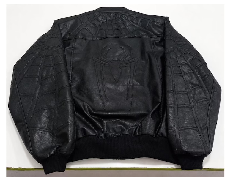 K&ouml;nnte beinhalten: Eine schwarze Bomberjacke mit einem strukturierten Spinnennetz-Muster, das auf die Schultern und &Auml;rmel gepr&auml;gt ist. Auf dem R&uuml;ckenteil befindet sich ein gro&szlig;es, zentriertes Spinnen-Emblem. Die Jacke hat einen gerippten Kragen, B&uuml;ndchen und Saum.
