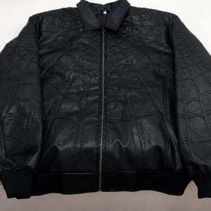 K&ouml;nnte beinhalten: Eine schwarze Jacke mit durchgehendem Frontrei&szlig;verschluss, klassischem Kragen und langen &Auml;rmeln mit gerippten B&uuml;ndchen. Die Au&szlig;enfl&auml;che ist mit einem erhabenen, gepr&auml;gten Spinnennetz-Muster verziert, das sich &uuml;ber Brust und &Auml;rmel erstreckt.