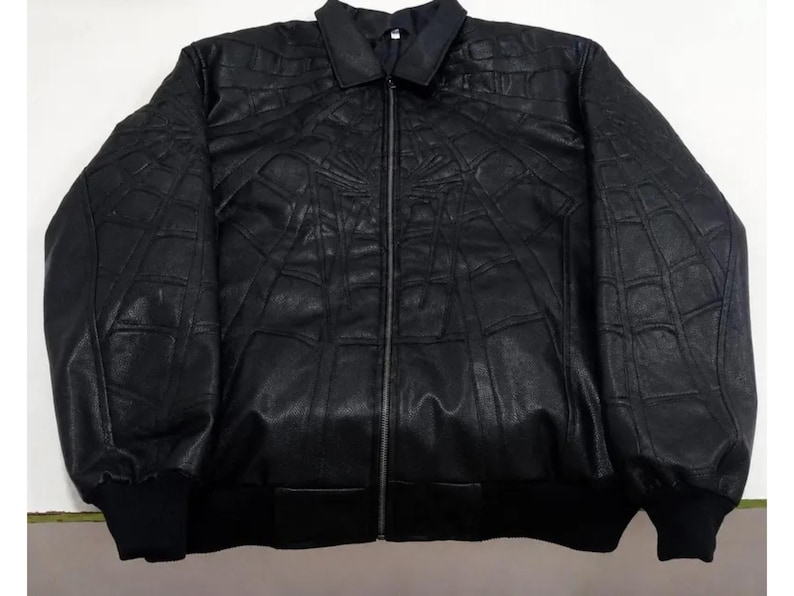 K&ouml;nnte beinhalten: Eine schwarze Jacke mit durchgehendem Frontrei&szlig;verschluss, klassischem Kragen und langen &Auml;rmeln mit gerippten B&uuml;ndchen. Die Au&szlig;enfl&auml;che ist mit einem erhabenen, gepr&auml;gten Spinnennetz-Muster verziert, das sich &uuml;ber Brust und &Auml;rmel erstreckt.