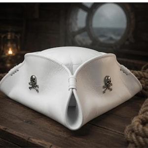 Op de afbeelding: Een witte driekante hoed van leer, voorzien van een gestructureerde rand met omgevouwen zijkanten. De hoed is versierd met twee metalen zilverkleurige emblemen van een schedel met gekruiste beenderen aan de voorzijde, vastgezet met zichtbare witte stiksels.